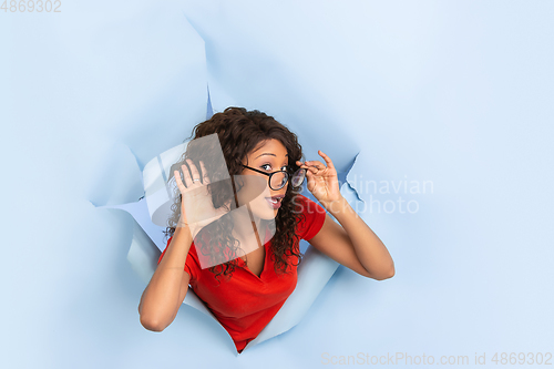 Image of Cheerful young woman poses in torn blue paper hole background, emotional and expressive