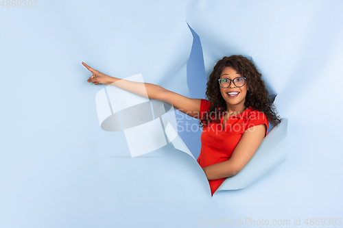 Image of Cheerful young woman poses in torn blue paper hole background, emotional and expressive