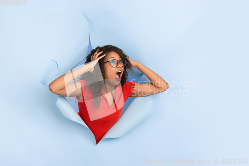 Image of Cheerful young woman poses in torn blue paper hole background, emotional and expressive