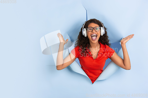 Image of Cheerful young woman poses in torn blue paper hole background, emotional and expressive