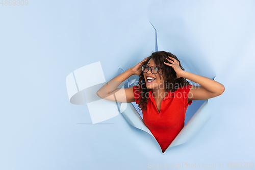 Image of Cheerful young woman poses in torn blue paper hole background, emotional and expressive