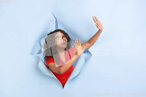 Image of Cheerful young woman poses in torn blue paper hole background, emotional and expressive