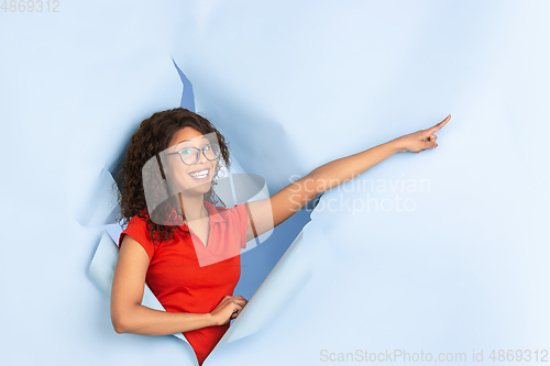 Image of Cheerful young woman poses in torn blue paper hole background, emotional and expressive