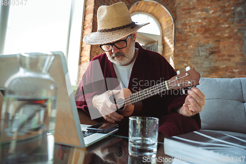 Image of Mature senior older man during quarantine, realizing how important stay at home during virus outbreak, giving concert of taking online lessons of guitar playing in Monte Carlo