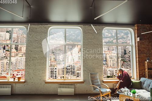 Image of Mature senior older man during quarantine, realizing how important stay at home during virus outbreak, looking throught window on day city Monaco