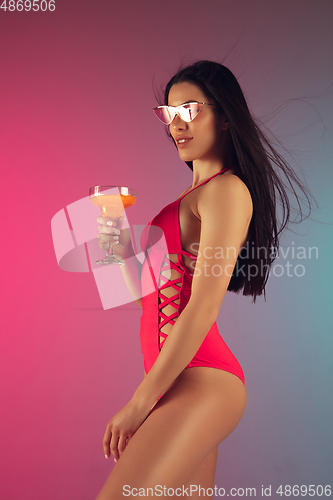 Image of Fashion portrait of young fit and sportive woman in stylish pink luxury swimwear with cocktail on gradient background. Perfect body ready for summertime.