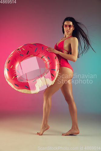 Image of Fashion portrait of young fit and sportive woman in stylish pink luxury swimwear with rubber donut on gradient background. Perfect body ready for summertime.