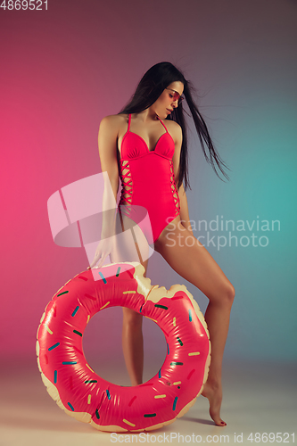 Image of Fashion portrait of young fit and sportive woman in stylish pink luxury swimwear with rubber donut on gradient background. Perfect body ready for summertime.