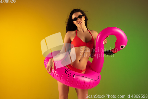 Image of Fashion portrait of young fit and sportive woman in stylish red luxury swimwear with rubber flamingo on gradient background. Perfect body ready for summertime.