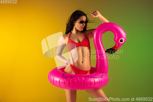 Image of Fashion portrait of young fit and sportive woman in stylish red luxury swimwear with rubber flamingo on gradient background. Perfect body ready for summertime.