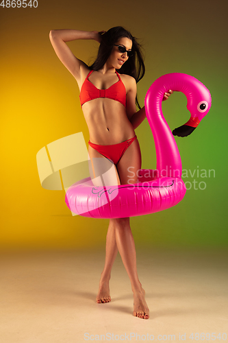 Image of Fashion portrait of young fit and sportive woman in stylish red luxury swimwear with rubber flamingo on gradient background. Perfect body ready for summertime.