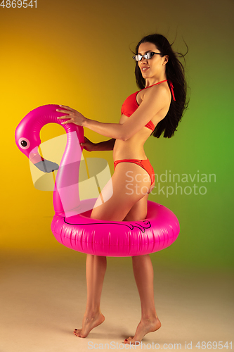 Image of Fashion portrait of young fit and sportive woman in stylish red luxury swimwear with rubber flamingo on gradient background. Perfect body ready for summertime.