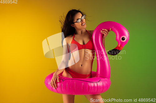 Image of Fashion portrait of young fit and sportive woman in stylish red luxury swimwear with rubber flamingo on gradient background. Perfect body ready for summertime.