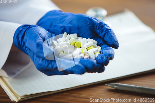 Image of Close up of doctors hands wearing blue protective gloves giving bunch of pills on wooden table background