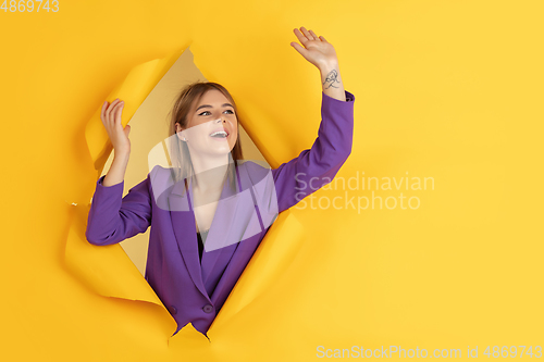 Image of Cheerful young woman poses in torn yellow paper hole background, emotional and expressive