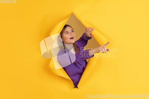 Image of Cheerful young woman poses in torn yellow paper hole background, emotional and expressive