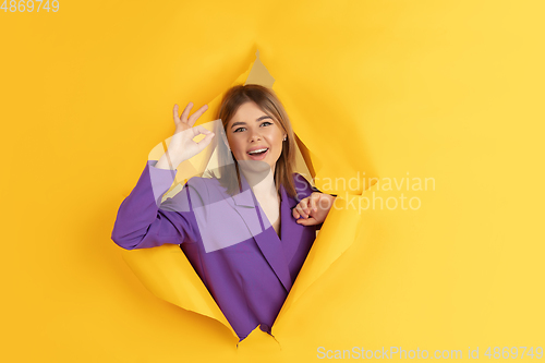 Image of Cheerful young woman poses in torn yellow paper hole background, emotional and expressive