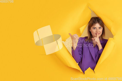 Image of Cheerful young woman poses in torn yellow paper hole background, emotional and expressive