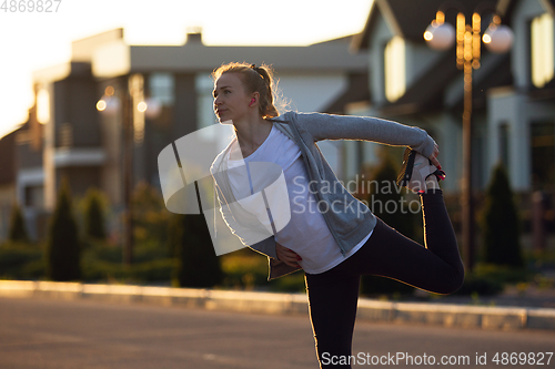 Image of Young female runner, athlete is stretching before jogging in the city street in sunshine. Beautiful caucasian woman training, listening to music