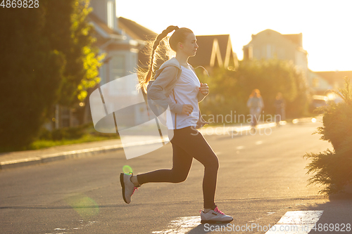 Image of Young female runner, athlete is jogging in the city street in sunshine. Beautiful caucasian woman training, listening to music