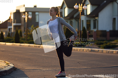 Image of Young female runner, athlete is stretching before jogging in the city street in sunshine. Beautiful caucasian woman training, listening to music