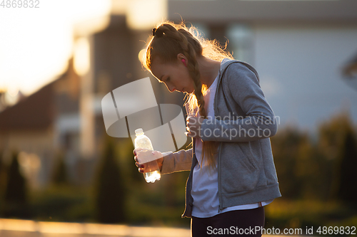 Image of Young female runner, athlete resting after jogging in the city street in sunshine. Beautiful caucasian woman training, listening to music