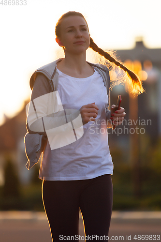 Image of Young female runner, athlete is jogging in the city street in sunshine. Beautiful caucasian woman training, listening to music