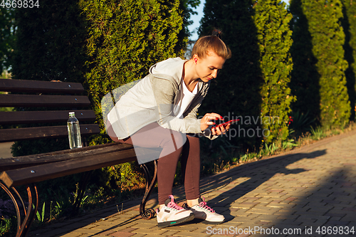 Image of Young female runner, athlete resting after jogging in the city street in sunshine. Beautiful caucasian woman training, listening to music