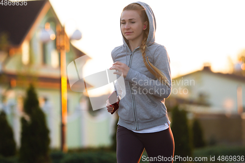 Image of Young female runner, athlete is jogging in the city street in sunshine. Beautiful caucasian woman training, listening to music