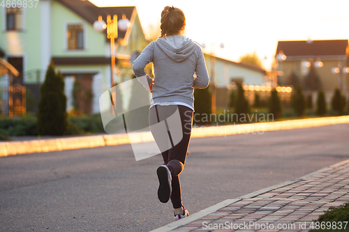 Image of Young female runner, athlete is jogging in the city street in sunshine. Beautiful caucasian woman training, listening to music