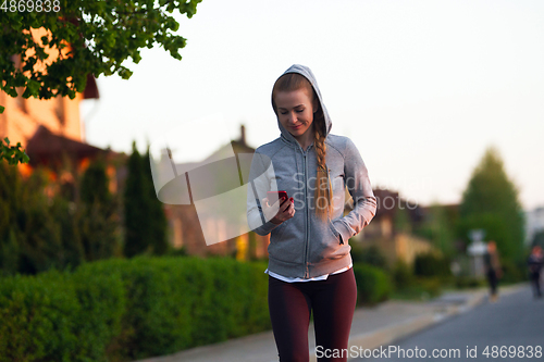 Image of Young female runner, athlete resting after jogging in the city street in sunshine. Beautiful caucasian woman training, listening to music