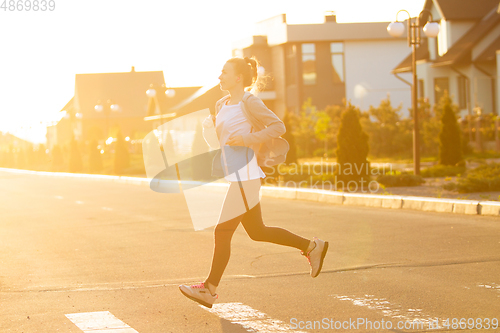 Image of Young female runner, athlete is jogging in the city street in sunshine. Beautiful caucasian woman training, listening to music