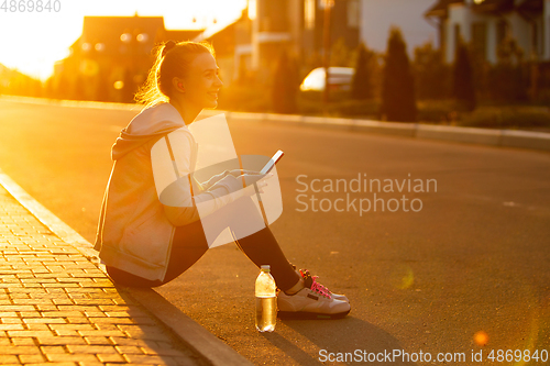 Image of Young female runner, athlete resting after jogging in the city street in sunshine. Beautiful caucasian woman training, listening to music