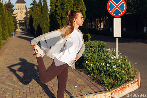 Image of Young female runner, athlete is stretching before jogging in the city street in sunshine. Beautiful caucasian woman training, listening to music