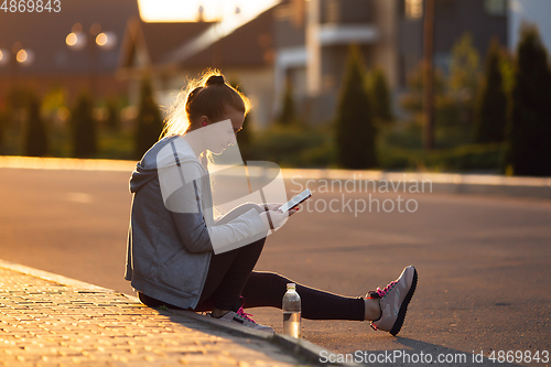 Image of Young female runner, athlete resting after jogging in the city street in sunshine. Beautiful caucasian woman training, listening to music