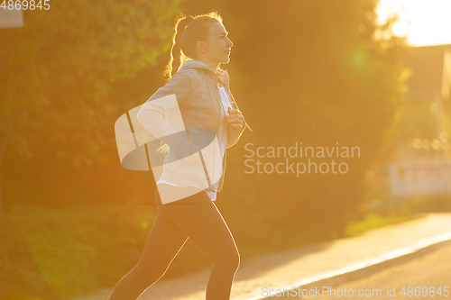 Image of Young female runner, athlete is jogging in the city street in sunshine. Beautiful caucasian woman training, listening to music