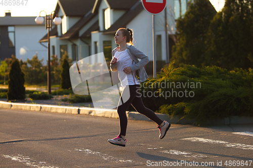 Image of Young female runner, athlete is jogging in the city street in sunshine. Beautiful caucasian woman training, listening to music