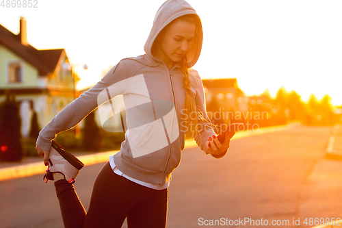 Image of Young female runner, athlete is stretching before jogging in the city street in sunshine. Beautiful caucasian woman training, listening to music