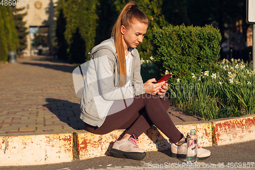 Image of Young female runner, athlete resting after jogging in the city street in sunshine. Beautiful caucasian woman training, listening to music