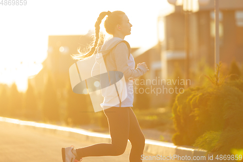 Image of Young female runner, athlete is jogging in the city street in sunshine. Beautiful caucasian woman training, listening to music