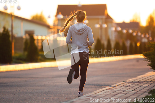 Image of Young female runner, athlete is jogging in the city street in sunshine. Beautiful caucasian woman training, listening to music