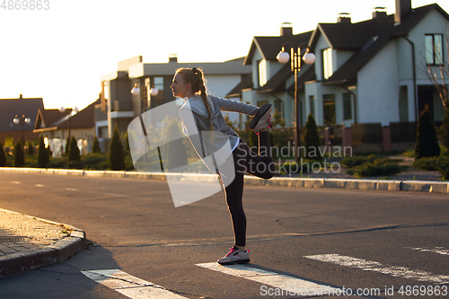 Image of Young female runner, athlete is stretching before jogging in the city street in sunshine. Beautiful caucasian woman training, listening to music