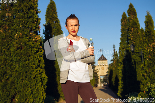 Image of Young female runner, athlete resting after jogging in the city street in sunshine. Beautiful caucasian woman training, listening to music