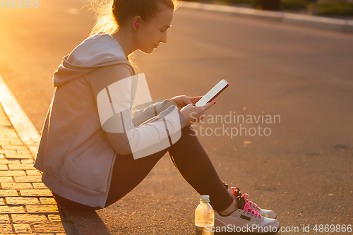 Image of Young female runner, athlete resting after jogging in the city street in sunshine. Beautiful caucasian woman training, listening to music