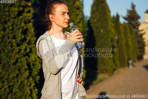 Image of Young female runner, athlete resting after jogging in the city street in sunshine. Beautiful caucasian woman training, listening to music