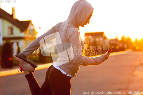 Image of Young female runner, athlete is stretching before jogging in the city street in sunshine. Beautiful caucasian woman training, listening to music