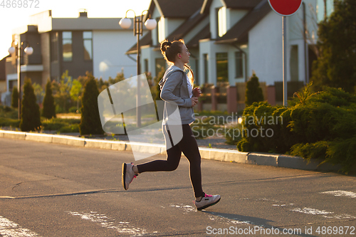 Image of Young female runner, athlete is jogging in the city street in sunshine. Beautiful caucasian woman training, listening to music