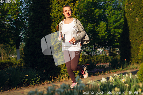 Image of Young female runner, athlete is jogging in the city street in sunshine. Beautiful caucasian woman training, listening to music