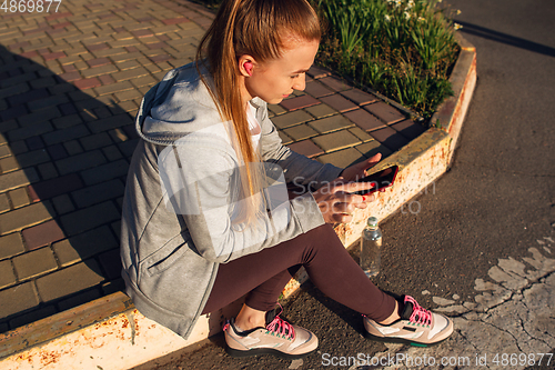 Image of Young female runner, athlete resting after jogging in the city street in sunshine. Beautiful caucasian woman training, listening to music