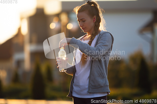 Image of Young female runner, athlete resting after jogging in the city street in sunshine. Beautiful caucasian woman training, listening to music